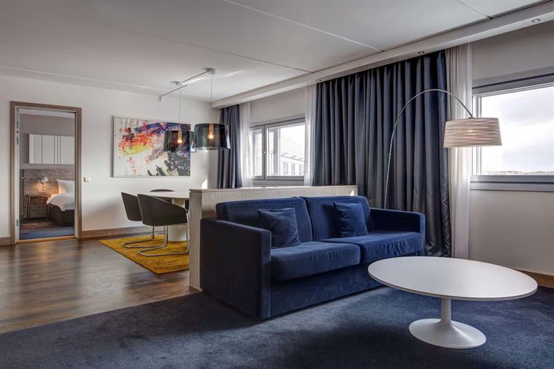 Interiør av suite med blå sofa og hvitt rundt bord, grå gardiner, hvitt spisebord og mørke stoler