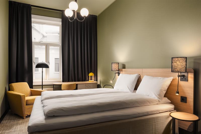 Et grønnmalt hotellrom med en dobbeltseng, en gul lenestol, et skrivebord og svarte gardiner
