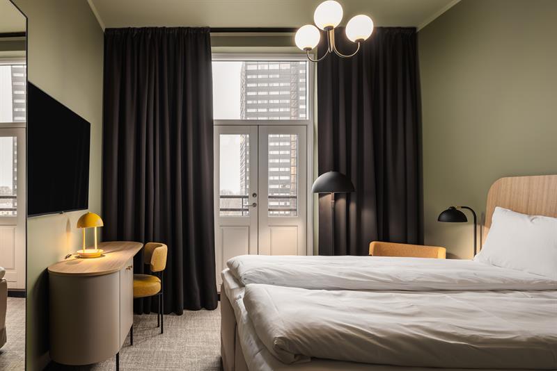 Et hotellrom med dobbeltseng, grønnmalte vegger, store svarte gardiner langs de store panoramavinduene og en TV på veggen