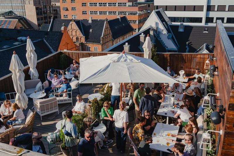 Takterrassen har sittegrupper, parasoller og grøntområder, med utsikt over de omkringliggende hustakene. Plassen er utformet for service og s