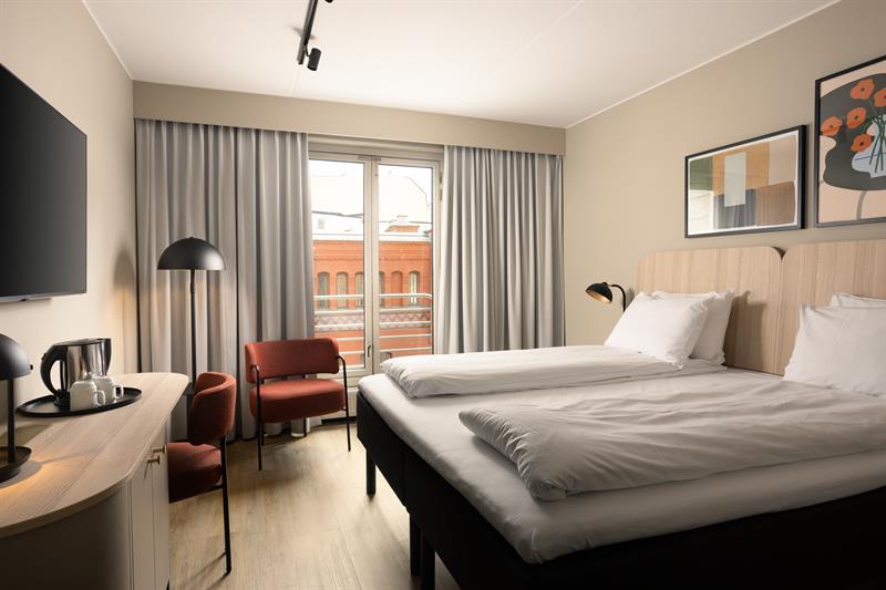 Hotellrom med stor dobbeltseng, TV på veggen, rød stol i hjørnet og grå gardiner