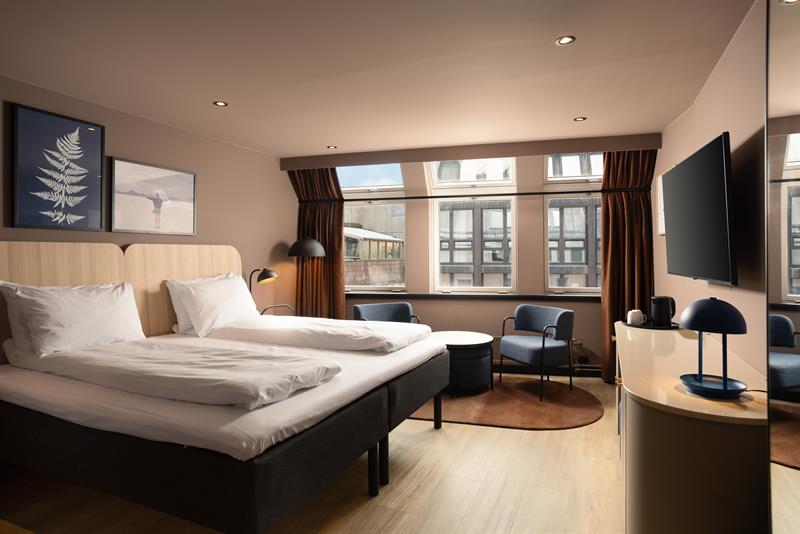 Hotellrom med stor dobbeltseng, TV på veggen, blå lenestoler og brune gardiner