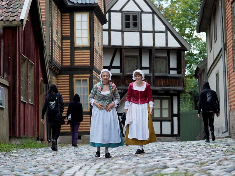 To kvinner i tradisjonelle klær går forbi gamle hus. Norsk Kulturhistorisk Museum