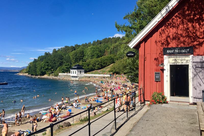 Et utsnitt mot stranden med folk som bader og soler seg, samt et rødt trehus. Skogen er synlig i bakgrunnen.