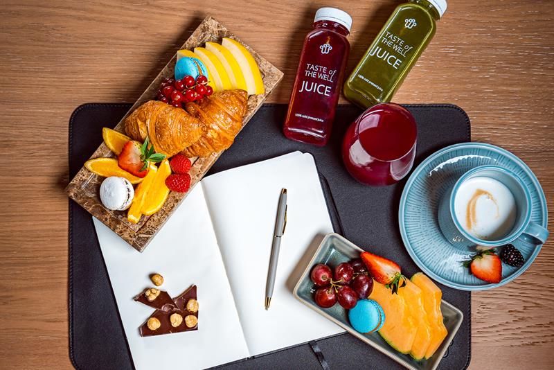 Frukt, en croissant, juiceflasker, et rødt glass, en blå kopp med cappuccino, en åpen notatblokk og en penn på et brunt trebord og en grå stol.