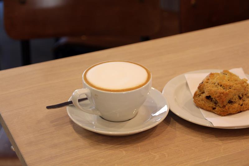 Detaljbilde av en kopp cappuccino og en scone på et bord.