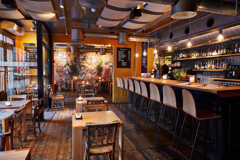 Restaurantinteriør med en bardisk og barstoler til høyre, firkantede trebord med stoler i midten. Gule vegger og tregulv.