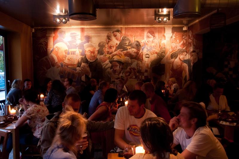 En fullsatt restaurant med folk som spiser og snakker sammen. Et stort maleri på veggen.