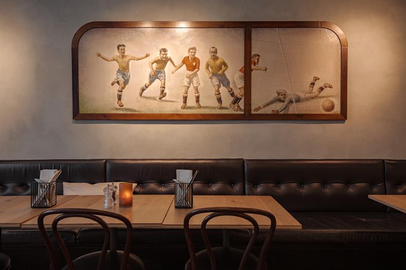 Et klassisk restaurantinteriør med polstrede benker, trebord og et veggmaleri av fotballspillere.