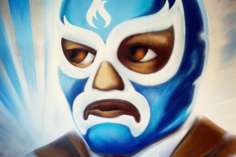 Detaljbilde av ulike matvarer, grønnsaker og frukt på trepinner foran et veggmaleri av en luchador.