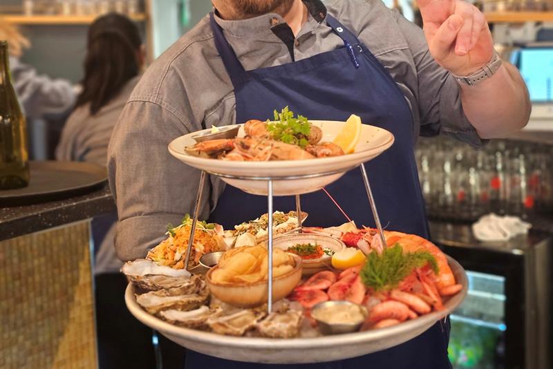 En restaurantansatt i uniform holder en stor sjømatfat og smiler mens han gjør et fredstegn
