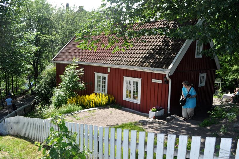 Et rødt trehus omgitt av trær, blomster og et hvitt stakittgjerde.