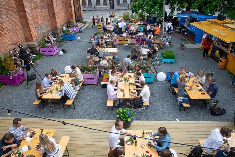 Utebilde fra en hagefest. Benker og bord med folk som drikker og snakker sammen.