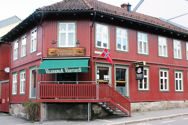 Utsiden av et rødt trehus med et norsk flagg hengende.