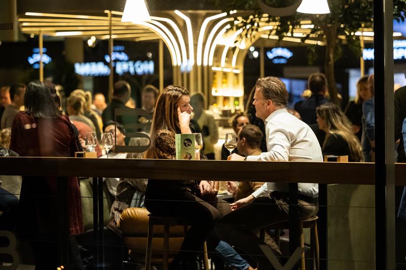 En kvinne og en mann sitter ved et bord og snakker sammen over et glass vin i en livlig og stemningsfull restaurant eller bar.
