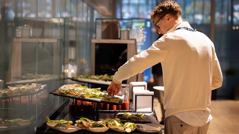 En mann med navneskilt tar seg noe fra en buffé med smørbrød og salat i et lyst konferanselokale.