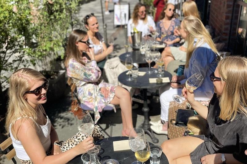 en gruppe kvinner som sitter i solen foran «Oh Dear» og skåler med vinglass 