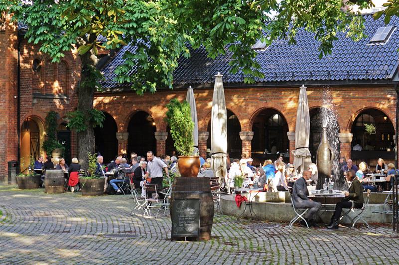 Uteservering med gjester som sitter, en murbygning i bakgrunnen.