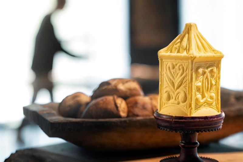 En utskåret figur av gult voks på en sokkel i forgrunnen, med brød og en uskarp figur i bakgrunnen.