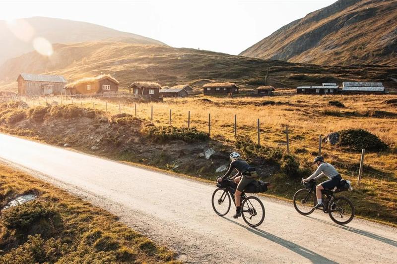 To personer som sykler langs en landevei i fjellet, med små hytter med torvtak i bakgrunnen.