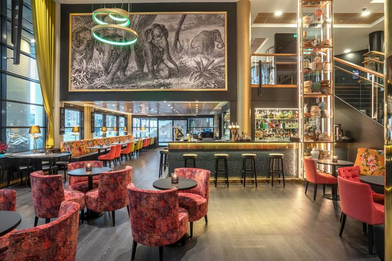 En bar med barkrakker, små runde bord med røde stoler, et stort veggmaleri av elefanter på veggen og en trapp i bakgrunnen