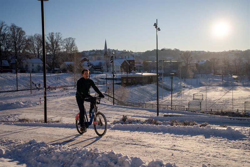 En mann som sykler langs en snødekt vei, med en fotballbane i bakgrunnen