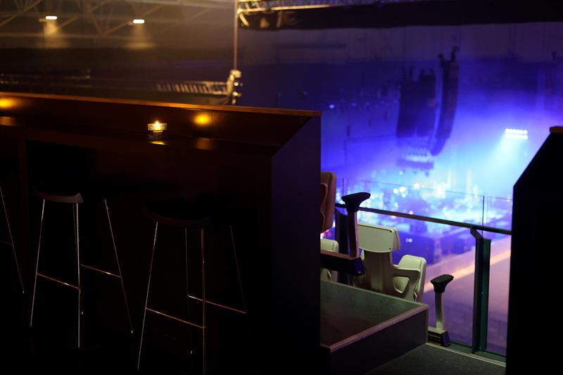 Utsikt fra en VIP-lounge i en konsertarena, med høye barstoler og tente stearinlys på disken i forgrunnen, og en scene opplyst av blått lys