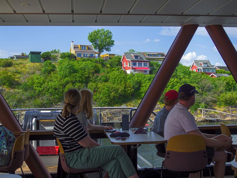 People sitting inside a boat looking out through the panoramic window at colourful houses and green landscape on the shore.