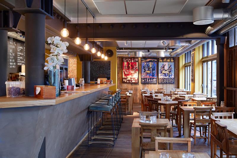 Restaurantinteriør med bardisk, barkrakker, mindre spisebord og stoler.