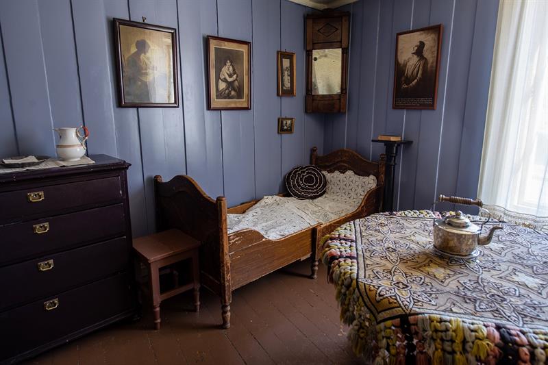 Historisk interiør med enkeltseng, kommode, bord og innrammede bilder på blåmalte vegger.