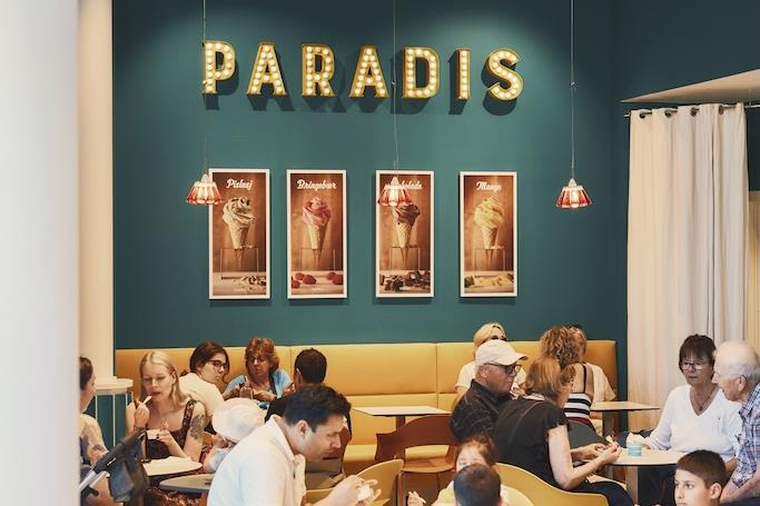 Folk sitter ved små, runde bord og spiser inne i kafeen. Paradis-logoen er på en grønn vegg på baksiden av lokalet.