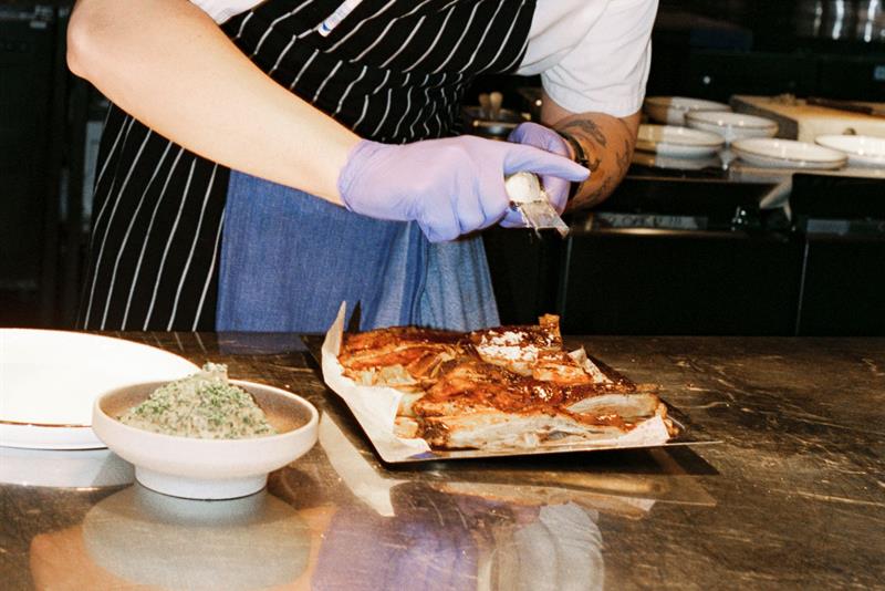 En kokk anretter kjøtt ved en kjøkkenbenk under varmt lys i et åpent restaurantkjøkken.