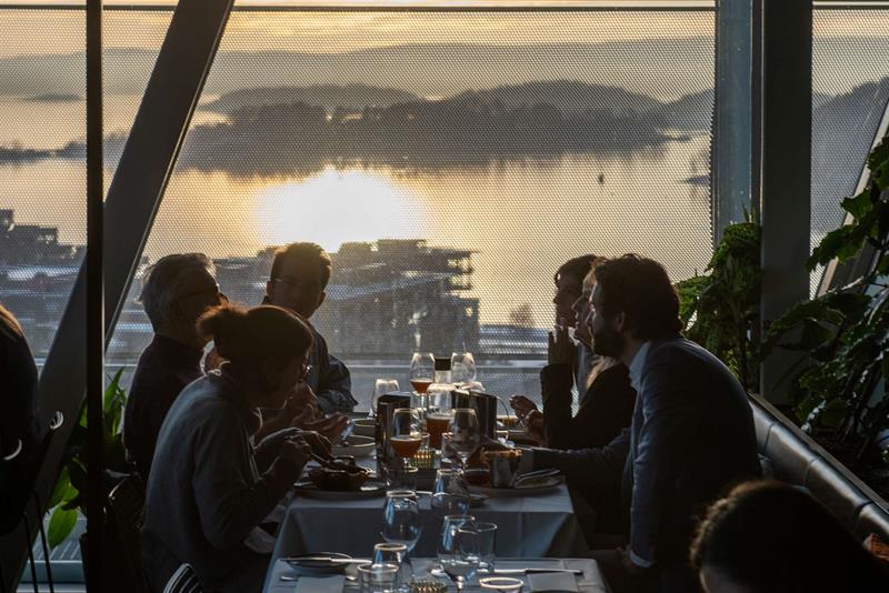 Folk nyter et måltid ved et langbord i en restaurant med utsikt over solnedgangen over Oslofjorden gjennom store vinduer