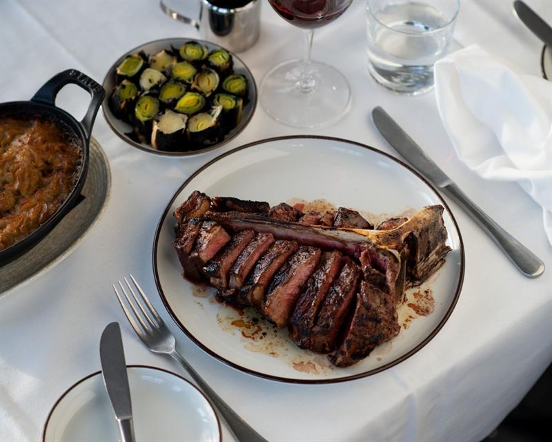 En grillet côte de boeuf servert på en hvit tallerken med tilbehør og vinglass på et dekket bord.
