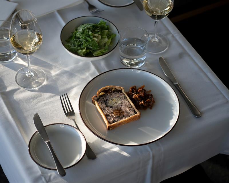 En tallerken med paté og tilbehør serveres på et elegant dekket bord i naturlig lys.