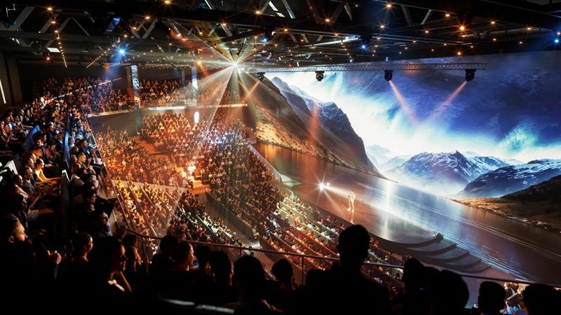 En fullsatt konferansesal med scene og storskjerm, der en foredragsholder presenterer for et stort publikum.