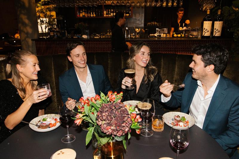 En gruppe på fire personer sitter rundt et restaurantbord og smiler og prater mens de nyter mat og drikke i en varm og stemningsfull atmosfære.