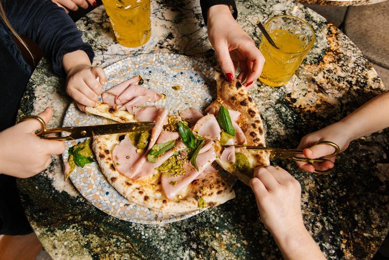 Flere hender deler en pizza med mortadella og pesto og klipper den med saks over et marmorbord.