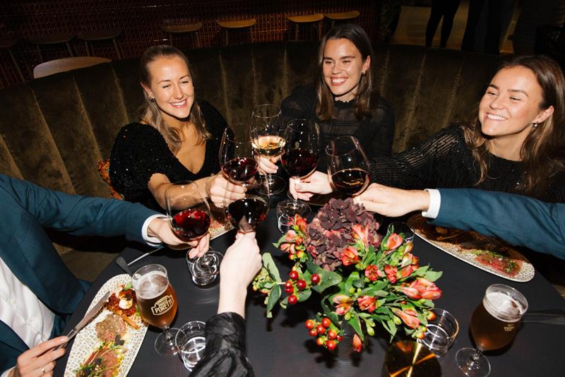 En gruppe mennesker skåler med vinglass over et restaurantbord, smilende og i feststemning.