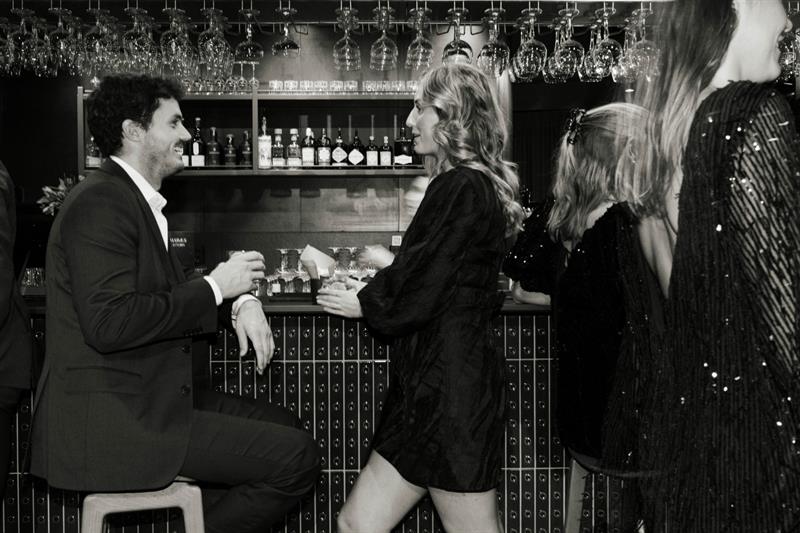 Folk kledd for en kveld ute samles i en bar, noen prater og andre går forbi, i en elegant og livlig setting.