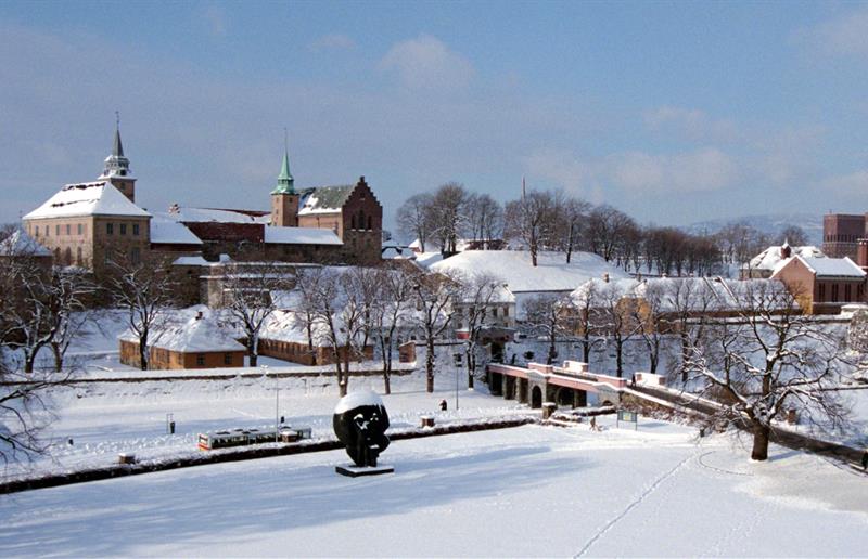 Vinterbilde av festningen dekket av snø, sett på lang avstand. Rådhuset i bakgrunnen på høyre side.