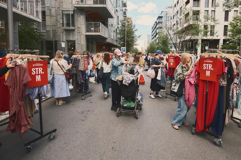 Folk kikker på klesstativer på et utendørs byttemarked omgitt av moderne boligblokker. Skiltene viser størrelser, og stemningen er livlig og så