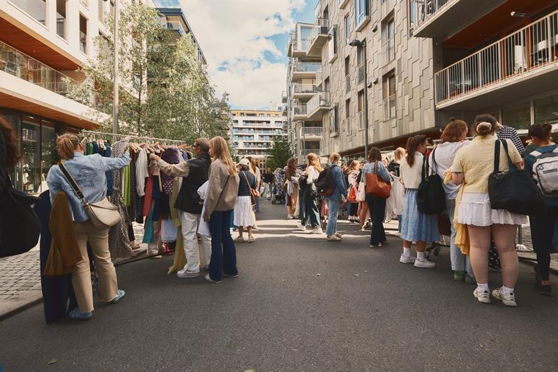 Folk kikker på klesstativer på et utendørs byttemarked omgitt av moderne boligblokker. Skiltene viser størrelser, og stemningen er livlig og så