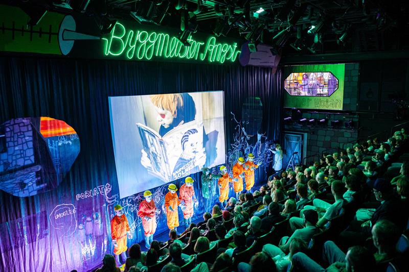 Sceneforestilling i et teater med skuespillere i fargerike kostymer foran et publikum.