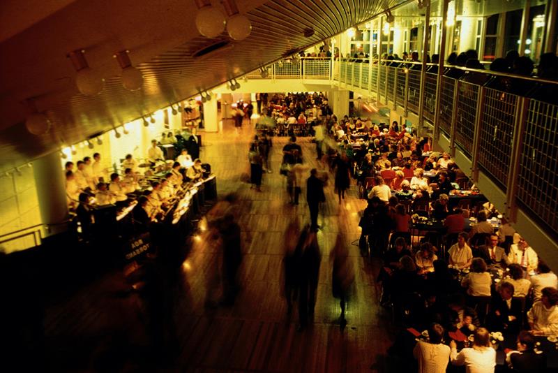 Stor teaterfoajé sett fra en balkong, med mange mennesker ved bordene og et orkester som spiller langs veggen.