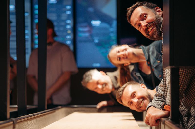 Smilende venner lener seg over et shuffleboardbord og følger med på pucken i et sosialt spillmiljø.