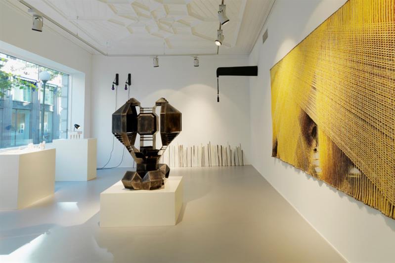 Galleriinteriør med et stort, gult, abstrakt maleri på høyre vegg og en skulptur i midten av rommet.