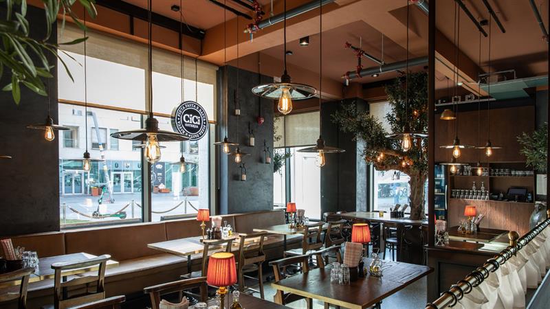 Restaurantinteriør med store vinduer, en brun skinnbenk, trestoler, bordlamper med rød skygge og industrielle hengelamper