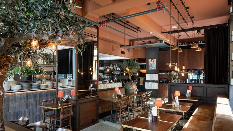 En restaurant med trebord, varme lamper, hengende lyspærer og et stort kunstig oliventre som sprer seg over sittegruppen