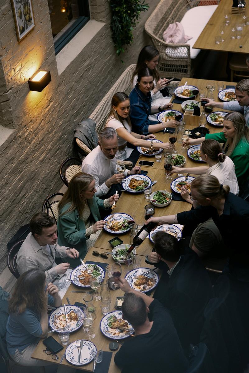 Gjestene sitter tett rundt et langbord og deler mat og vin i en livlig, sosial restaurantatmosfære sett ovenfra.